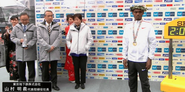 東京マラソン表彰式でポケットに手を入れ傘をさしてもらっている小池都知事の画像